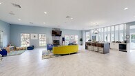 Located at 7595 Baymeadows Cir W in Jacksonville, FL, this single-story community clubhouse offers a well-rounded collection of amenity spaces designed for relaxation, recreation, and productivity. Throughout the facility, soft blue-gray walls, marble-look tile flooring, and recessed lighting create a clean, modern aesthetic that remains consistent from room to room.

The central lounge serves as the heart of the clubhouse, featuring a bold yellow curved sofa, blue accent chairs, a marble-surround fireplace with a wall-mounted TV, and a bar-height counter with seating — all bathed in natural light from multiple windows. An adjacent dining and coworking area provides round tables, upholstered chairs, a whiteboard, and greenery, making it equally suited for casual meals or focused work sessions.

A dedicated children's play area with colorful rugs and age-appropriate furnishings sits alongside the main lounge, offering a comfortable space for younger residents. The game and media room features a pool table, a wall-mounted TV with modular lounge seating in yellow and blue, and computer workstations with direct views of the outdoor pool area.

The fitness facilities are divided into two rooms: a cardio and strength training room equipped with treadmills and cable machines, and a separate functional training studio with a power rack, punching bag, battle ropes, and free weights. Together, these spaces deliver a comprehensive, amenity-rich environment for community living.