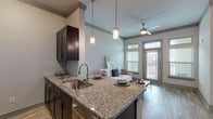 This single-floor residential unit offers a thoughtfully designed open-concept layout that seamlessly connects the kitchen, dining, and living areas. The kitchen is a standout feature, equipped with sleek dark cabinetry, stainless steel appliances including a full-size refrigerator, microwave, and range, complemented by granite countertops and a peninsula island with bar seating. Pendant lighting and wood-look flooring throughout the main living area add warmth and style. The living room is bright and welcoming, featuring elevated ceilings, large windows that invite generous natural light, and a glass-paneled door leading to an outdoor balcony. A ceiling fan overhead enhances comfort in the living space. The bedroom is a calm retreat, finished with plush carpeting, expansive windows offering outdoor views, and ample natural light. The bathroom is well-appointed with granite vanity counters, dark cabinetry consistent with the kitchen aesthetic, a full soaking tub, and direct access to a spacious walk-in closet with built-in shelving and hanging rods. Crown molding throughout the unit adds a refined architectural touch, tying together the cohesive and contemporary design of the space.