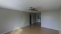This single-story space presents an open, straightforward layout with a functional and practical design. The main living area is finished with tile flooring throughout, offering durability and ease of maintenance. Neutral gray walls create a clean, understated backdrop, while white baseboards provide a crisp contrast along the perimeter of the room.

A ceiling fan with dark wood-toned blades and a brushed nickel finish is centrally mounted, serving both a decorative and practical purpose for air circulation. The textured popcorn ceiling adds dimension overhead. Natural light enters through unseen windows, casting bright patterns across the tile floor.

Visible from the main living area, the kitchen occupies a compact space toward the rear of the unit. White appliances, including what appears to be a refrigerator, are visible, along with a light-colored countertop. A closet or storage area is also accessible nearby, adding to the functional storage capacity of the space.

The open sightlines between the living area and kitchen create a connected flow, making the overall layout feel cohesive. The space presents a blank canvas, well-suited for a variety of furnishing arrangements.