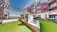 This well-appointed outdoor courtyard is set within a striking multi-story residential building that blends classic red brick with modern architectural elements. The courtyard is thoughtfully designed to offer residents a resort-like outdoor experience, featuring a combination of vibrant artificial turf lawn areas, elegant stone-tiled walkways, and raised brick planters filled with lush coastal greenery and ornamental grasses.

A standout centerpiece of the space is a linear fire feature framed in brick and filled with dark decorative rock, providing a sophisticated focal point for outdoor gatherings. White-painted pergolas span across multiple zones of the courtyard, creating shaded retreats while adding architectural interest overhead. Comfortable Adirondack chairs are positioned throughout the turf areas, inviting relaxation and social interaction.

Lush landscaping, including a prominent palm tree and a variety of tropical and ornamental plantings, reinforces the coastal character of the space. A herringbone-patterned brick accent wall adds a decorative touch and privacy to one corner of the courtyard.

Framed by the building on multiple sides, the courtyard opens toward sweeping ocean views, allowing natural light and sea breezes to flow through the space. The overall ambiance is welcoming, polished, and ideal for outdoor living.