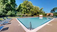Located at 402 Westover Hills Blvd in Richmond, Virginia, this outdoor recreation area offers a well-appointed community amenity space designed for relaxation and leisure. The centerpiece is a generously sized swimming pool, surrounded by a wide concrete sun deck lined with blue lounge chairs, providing ample space for sunbathing and poolside enjoyment. An adjacent brick utility building with restroom facilities serves the pool area conveniently. A standout feature of the grounds is a wooden pergola structure that offers shaded outdoor seating, furnished with tables and chairs — ideal for gathering and socializing. Raised brick planter beds add decorative greenery throughout the space, while mature trees along the perimeter provide natural shade and a lush backdrop. The entire area is enclosed by a sturdy black wrought iron fence, ensuring security and a clean, defined boundary. Expansive, well-maintained lawns extend across the grounds, creating open green spaces that complement the hardscaped areas. Lighting poles are positioned throughout for evening use. The space benefits from an open sky setting with abundant natural light, making it an inviting outdoor destination throughout the warmer months.