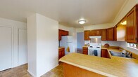 This single-floor residence offers a straightforward, functional layout designed for comfortable everyday living. The kitchen is a standout space, featuring warm honey-toned wood cabinetry that extends from floor to ceiling, providing generous storage. Light wood-toned countertops run along the perimeter and wrap around a central peninsula, offering ample prep space and a natural divider between the kitchen and the adjoining living area. White appliances, including a range with overhead ventilation and a refrigerator, are neatly integrated into the cabinetry. A window above the double-basin sink allows natural light to fill the room and provides a view to the outdoors. The living area is open and spacious, finished with neutral wall-to-wall carpeting that continues throughout the bedroom spaces. Each bedroom is generously proportioned with clean white walls, carpeted floors, and windows that invite natural light. One bedroom includes a closet and direct hallway access. The bathroom features a full tub-shower combination with tiled flooring, a vanity with a marble-style countertop, a mirrored medicine cabinet, and clean white finishes throughout. The overall aesthetic of the space is clean and neutral, offering a versatile canvas for personalization.