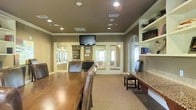 This single-floor space is designed to accommodate both professional and communal needs, centered around a formal meeting or conference area. The main room features rich hardwood flooring that transitions to carpeted zones, creating a warm and inviting atmosphere. A large wooden conference table surrounded by leather chairs serves as the focal point, complemented by a wall-mounted flat-screen television overhead. Recessed lighting is evenly distributed across the taupe-toned ceiling, accented by elegant crown molding that adds a refined architectural touch. Built-in open shelving along one wall provides organized display and storage space, while a granite-topped counter adds a practical and polished surface. French-style glass-paned doors lead to adjacent rooms and offices, allowing natural light to filter throughout and maintaining an open, connected feel. Additional interior windows and glass partitions further enhance the sense of transparency and accessibility. The neutral earth-tone color palette throughout creates a professional yet comfortable environment, making this space well-suited for meetings, collaborative work, or community gatherings.