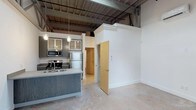 Located at 1200 S 2nd St in Wilmington, NC, this single-floor space showcases a compelling blend of industrial and contemporary design. The open layout is anchored by soaring ceilings with exposed structural beams, steel joists, and visible ductwork, lending the space a distinctly urban, loft-like character. Polished concrete floors run throughout, adding to the industrial aesthetic while remaining easy to maintain.

The kitchen is a standout feature, equipped with stainless steel appliances including a range, microwave, and refrigerator. Sleek gray wood cabinetry with glass-front upper cabinets provides ample storage, while the kitchen island with an integrated sink serves as a functional centerpiece. Pendant lighting with an Edison-style design hangs above, offering warm, focused illumination and adding a stylish finishing touch.

Decorative column molding frames the entryway to an adjoining room or hallway, introducing a classic architectural contrast against the raw industrial backdrop. The crisp white walls and generous vertical space allow natural and ambient light to fill the interior, creating a bright and welcoming atmosphere. The result is a thoughtfully designed space that balances raw, warehouse-inspired elements with modern residential finishes.