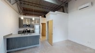 Located at 1200 S 2nd St in Wilmington, NC, this single-floor space showcases a compelling blend of industrial and contemporary design. The open layout is anchored by soaring ceilings with exposed structural beams, steel joists, and visible ductwork, lending the space a distinctly urban, loft-like character. Polished concrete floors run throughout, adding to the industrial aesthetic while remaining easy to maintain.

The kitchen is a standout feature, equipped with stainless steel appliances including a range, microwave, and refrigerator. Sleek gray wood cabinetry with glass-front upper cabinets provides ample storage, while the kitchen island with an integrated sink serves as a functional centerpiece. Pendant lighting with an Edison-style design hangs above, offering warm, focused illumination and adding a stylish finishing touch.

Decorative column molding frames the entryway to an adjoining room or hallway, introducing a classic architectural contrast against the raw industrial backdrop. The crisp white walls and generous vertical space allow natural and ambient light to fill the interior, creating a bright and welcoming atmosphere. The result is a thoughtfully designed space that balances raw, warehouse-inspired elements with modern residential finishes.