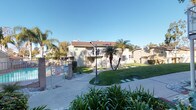 This residential community presents a welcoming and well-kept exterior environment characterized by its classic architectural style. The buildings feature neutral-toned horizontal lap siding, dark brown pitched rooflines, and white-trimmed windows, giving the complex a cohesive and tidy appearance. Exterior stairways with metal railings provide access between levels, and each building is thoughtfully positioned to create open, airy common areas in between.

The grounds are a standout feature of this community, with expansive stretches of neatly manicured green lawn running between the buildings. Mature palm trees and towering eucalyptus trees line the property, adding natural shade and a distinctly warm, Southern California character. Ornamental black lamp posts line the central walkways, enhancing both the aesthetic and the functionality of the pedestrian paths that wind throughout the complex.

A community pool area is visible along the perimeter, enclosed by a fence and surrounded by additional tropical landscaping, including tall royal palms. Lush shrubs, ground cover plantings, and seasonal greenery further enrich the landscape, contributing to a vibrant and pleasant outdoor atmosphere. The entire complex benefits from abundant natural sunlight under clear blue skies, reinforcing the bright, open, and inviting ambiance of this well-established residential community.