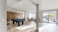 This well-appointed amenity space spans a generous single floor, offering a thoughtfully designed layout that blends recreation, relaxation, and functionality. The open-concept living area is anchored by light-toned hardwood-style flooring that runs throughout, complementing the clean, contemporary aesthetic defined by crisp white walls and recessed lighting.

The games zone is a standout feature, centering around a full-size pool table set against a warm-toned accent wall adorned with a decorative sunburst mirror and artwork. This area flows seamlessly into a lounge space furnished with mid-century modern armchairs and a sofa, bathed in natural light from large windows and sliding glass doors that open to an outdoor patio.

A separate seating nook near the windows provides a quieter retreat, fitted with a wall-mounted air conditioning unit for year-round comfort. The dining area features a rectangular table with stylish upholstered chairs, positioned adjacent to a compact kitchenette equipped with stainless steel appliances, including a refrigerator and microwave, set within white cabinetry.

The accessible bathroom is finished with grey subway tile, stainless steel grab bars, and matching hardwood-style flooring, ensuring both style and practicality. Vaulted ceiling sections add a sense of openness and volume, while the abundant glazing throughout connects the interior to the surrounding landscaped grounds visible from the exterior.