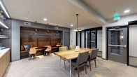 This thoughtfully designed single-floor space blends contemporary aesthetics with functional communal gathering areas. The interior is anchored by a striking live-edge wood dining table surrounded by upholstered gray chairs, serving as the centerpiece of the main gathering area. Overhead, a cluster of modern gold pendant lights adds warmth and visual interest to the space.

Adjacent to the dining area is a stylish banquette seating nook, featuring richly tufted caramel leather booth seating set against a textured reclaimed wood accent wall. Copper-toned pendant lights illuminate this cozy corner, which is complemented by light wood bistro-style chairs and small round tables, creating an inviting lounge atmosphere.

The flooring throughout consists of large-format herringbone-patterned tile in a cool gray tone, lending a refined, contemporary feel underfoot. The ceiling is finished in crisp white with recessed lighting supplementing the decorative pendants for well-distributed illumination.

On one side of the space, sleek black-framed glass partition walls define a separate enclosed area, adding a modern industrial touch while maintaining visual openness. The overall color palette—combining warm wood tones, gray upholstery, and cool tile—creates a balanced and welcoming environment suited for both casual gatherings and productive meetings.