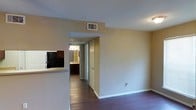 This single-floor space features an open-concept layout that connects the main living area to the kitchen and beyond. The living area is spacious and open, with warm beige walls and dark hardwood flooring that runs throughout, providing a cohesive and grounded aesthetic. A partial wall with a countertop passthrough separates the living area from the kitchen, which is visible to the left and includes dark cabinetry and a black refrigerator. This design maintains an open feel while defining each functional zone. A central hallway extends from the main living space, leading to additional rooms at the rear of the unit. A door is visible at the end of the hallway, and a well-lit room—likely a bathroom—can be seen branching off, illuminated by a chandelier-style light fixture. The ceilings are standard height and include a centrally located HVAC vent, indicating a forced-air climate control system. White baseboards and door trim provide clean contrast against the darker flooring and neutral walls. Overall, the space offers a practical and straightforward layout with a functional flow between living, kitchen, and private areas.