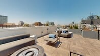 Located in the heart of Los Angeles, this rooftop terrace at 3920 W 5th St offers a striking outdoor retreat above the city. The space is finished with large-format wood-look porcelain tile laid in a herringbone and linear pattern, lending a refined, contemporary aesthetic throughout. Generous raised planters constructed in smooth concrete house a curated mix of drought-tolerant succulents, agaves, ornamental grasses, and colorful accent plants, adding vibrant greenery and texture to the open-air environment. A dedicated lounge area is furnished with modern modular seating, armchairs with blue accent cushions, and a fire pit, creating an inviting setting for gatherings and relaxation. The terrace offers sweeping, unobstructed panoramic views in multiple directions — from the iconic Los Angeles skyline and mid-rise commercial towers to the east, to tall palm trees and the distant San Gabriel Mountains to the north and west. Metal railing sections define the perimeter and frame the vistas without interrupting sight lines. The rooftop also features utility infrastructure including HVAC equipment and stairwell access points, neatly integrated into the overall design. Under the clear Southern California sky, this rooftop terrace delivers an exceptional urban outdoor experience.