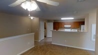 This single-floor residence offers a practical and comfortable open-concept layout that seamlessly connects the living area to the kitchen. The living space features neutral-toned walls, soft carpeted flooring, and a ceiling fan with integrated lighting that anchors the room. Clean white baseboards and trim throughout provide a crisp contrast to the warm beige walls.

The kitchen is positioned adjacent to the living area and is defined by a peninsula-style breakfast bar, providing both a natural division of space and additional seating or prep surface. Warm medium-toned wood cabinetry lines the upper walls of the kitchen, offering ample storage, while white appliances — including a range and microwave — are neatly integrated beneath. Fluorescent overhead lighting ensures the kitchen remains bright and functional.

A hallway visible from the main living area leads to additional rooms, hinting at a well-organized floor plan beyond the open common area. The laundry area, partially visible through a doorway, suggests in-unit conveniences. The overall ambiance of the space is warm and understated, with a neutral color palette that provides a versatile backdrop suited to a variety of interior styles. The layout is efficient, making excellent use of the available space throughout.