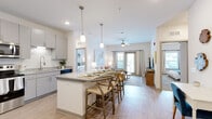 This well-appointed single-floor residence showcases a bright, open-concept layout that seamlessly connects the kitchen, dining, and living areas. The kitchen is outfitted with white shaker-style upper cabinets, gray lower cabinetry, granite countertops, a white subway tile backsplash, and stainless steel appliances including a refrigerator, range, and microwave. A spacious island with pendant lighting above serves as both a prep area and casual dining space with bar seating. Light wood-style flooring runs throughout the main living areas, enhancing the airy, contemporary feel. The living room features a ceiling fan, recessed lighting, and access to an outdoor area through glass doors that invite natural light. Two bedrooms are finished with plush carpet, multiple windows with blinds and curtain panels, and ceiling fans for comfort. Each bedroom is generously sized and accompanied by a well-designed bathroom featuring granite-topped vanities, large framed mirrors, and wood-look flooring. One bathroom includes walk-in closet access with wire shelving. A walk-in closet with wire rack storage is also available as dedicated storage. The overall aesthetic is clean and modern with a neutral palette throughout.
