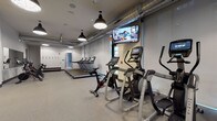 Located at 101–165 E Louisiana St in downtown Indianapolis, this well-appointed fitness center offers a comprehensive workout environment designed for a variety of exercise routines. The space is organized into distinct functional zones, allowing users to move seamlessly between cardio, strength training, and cycling areas.

The cardio section features a row of Matrix treadmills and multiple elliptical cross-trainers, each equipped with individual digital screens. Peloton and spin-style cycling bikes occupy their own dedicated area alongside a mirrored wall, creating a studio-like atmosphere for cycling workouts. A wall-mounted flat-screen TV provides additional entertainment throughout the space.

The strength and functional training zone is outfitted with a power rack, TRX suspension trainers, medicine balls, stability balls, and a rack of dumbbells — offering a full range of resistance and functional fitness options. A built-in storage unit provides organized space for accessories and personal items.

The industrial-inspired design is highlighted by exposed ductwork running along the ceiling, large ceiling fans, and bold pendant lighting with black fixtures. Accent walls in yellow and dark charcoal add visual contrast against the neutral gray tones throughout. Durable rubber flooring covers the entire space, and large windows allow natural light to filter in, enhancing the open, energetic ambiance of this modern fitness facility.