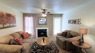 Located in Amarillo, Texas, this single-floor home offers a comfortable and welcoming living environment. The living room serves as the heart of the space, centered around a classic fireplace accented by a decorative mirror above the mantle, creating a warm and inviting focal point. A ceiling fan with integrated lighting keeps the room comfortable year-round while adding a practical touch to the layout. Plush seating, including two sofas arranged around a wooden coffee table, provides ample space for relaxing or entertaining. Natural light filters in through windows dressed with patterned curtains, brightening the carpeted floors and adding a soft, homey atmosphere. A textured ceiling adds subtle visual interest overhead. The layout flows naturally, with an entry door visible from the living area, suggesting easy access to the rest of the home. The overall ambiance is warm and lived-in, combining functional furnishings with personal touches that give the space a cozy, residential character well-suited to everyday comfort in the heart of the Texas Panhandle.