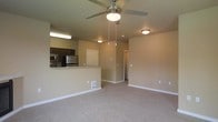 This single-floor residence offers an open-concept layout that seamlessly connects the living area and kitchen, creating a functional and cohesive living environment. The living area is finished with wall-to-wall carpeting in a neutral beige tone, contributing to a warm and inviting atmosphere. A ceiling fan with a built-in light fixture is centrally positioned overhead, providing both air circulation and ambient lighting to the space.

The kitchen is designed with dark-toned cabinetry that contrasts neatly against the light-colored countertops, which extend to form a raised breakfast bar — an ideal feature for casual dining or additional workspace. Stainless steel appliances, including a refrigerator and microwave, are visible and add a modern, functional touch to the cooking area.

The walls throughout are finished in a soft, neutral beige, creating a cohesive and understated backdrop that complements the overall design. Flush-mount ceiling lights provide consistent illumination throughout the space. A hallway visible from the main living area leads to additional rooms, suggesting a practical and well-organized floor plan. Overall, the space presents a clean, move-in-ready environment with a straightforward, modern aesthetic.
