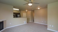 This single-floor residence offers an open-concept layout that seamlessly connects the living area and kitchen, creating a functional and cohesive living environment. The living area is finished with wall-to-wall carpeting in a neutral beige tone, contributing to a warm and inviting atmosphere. A ceiling fan with a built-in light fixture is centrally positioned overhead, providing both air circulation and ambient lighting to the space.

The kitchen is designed with dark-toned cabinetry that contrasts neatly against the light-colored countertops, which extend to form a raised breakfast bar — an ideal feature for casual dining or additional workspace. Stainless steel appliances, including a refrigerator and microwave, are visible and add a modern, functional touch to the cooking area.

The walls throughout are finished in a soft, neutral beige, creating a cohesive and understated backdrop that complements the overall design. Flush-mount ceiling lights provide consistent illumination throughout the space. A hallway visible from the main living area leads to additional rooms, suggesting a practical and well-organized floor plan. Overall, the space presents a clean, move-in-ready environment with a straightforward, modern aesthetic.