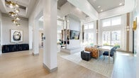 This thoughtfully designed single-floor communal space presents a sophisticated blend of modern aesthetics and functional design. The hallway entry sets an immediate tone with warm oak hardwood flooring, structural columns, globe pendant lighting in brushed gold, and a striking geometric mirror installation that adds depth and visual intrigue.

The main lounge area is defined by soaring double-height ceilings that amplify the sense of openness and volume. Oversized windows line the perimeter, inviting abundant natural light and offering views of the surrounding exterior landscape. The lounge is furnished with a tufted caramel leather sofa, organic sculptural white accent chairs, and a textured stone cube coffee table atop a layered area rug. A wall-mounted flat-screen television and elongated pendant light fixtures anchor the space, while a built-in work table with upholstered seating provides a collaborative area.

An eye-catching teal and white geometric tile accent wall near the entrance adds a bold artistic focal point, complemented by sculptural gold wall sconces. The kitchen features a generous waterfall-edge white quartz island with bar stool seating, warm wood cabinetry, open shelving against a patterned tile backsplash, and an organic branching chandelier overhead. Natural light floods in through a large window, completing this well-curated, multi-functional space.