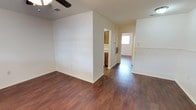 This single-story space showcases a straightforward, functional layout with a clean and neutral aesthetic throughout. The main living area is finished with rich, dark hardwood-style flooring that extends seamlessly into the adjoining hallway and kitchen, creating a cohesive visual flow across the open floor plan. White walls and white trim provide a bright, clean backdrop that enhances the sense of openness throughout the space.

A ceiling fan is installed in the main living area, contributing to air circulation and comfort. Overhead vents are visible, indicating a central HVAC system. The layout features a partial wall that subtly separates the living area from the kitchen corridor, adding a degree of definition without closing off the space.

The kitchen, visible through a passthrough opening, includes warm wood-toned cabinetry and continues the hardwood-style flooring. A hallway leading toward a naturally lit doorway suggests a direct entry point, likely a front or side entrance, which introduces natural light into the interior. The overall ambiance is tidy and move-in ready, with a neutral palette that allows for versatile furnishing and décor options.