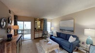 This single-floor residence centers around a well-appointed living area designed for both comfort and functionality. The living room features soft, wall-to-wall carpeting and is bathed in natural light streaming through a large window and sliding glass doors that open onto a private balcony, where outdoor seating is visible. A textured popcorn ceiling adds a classic touch overhead, while warm beige walls create a neutral, inviting backdrop throughout the space. The living area comfortably accommodates a full-sized sofa and ottoman arrangement, anchored by a patterned area rug. A large landscape-style artwork adorns one wall, reinforcing the coastal ambiance of the room. Adjacent to the main living area, a built-in vanity or wet bar with wood cabinetry and a decorative mirror adds a distinctive architectural feature, providing both storage and visual interest. Patterned drapes frame the sliding door, adding depth and texture. The overall layout flows openly, with the indoor and outdoor areas connecting seamlessly to enhance the sense of space and provide an airy, relaxed atmosphere throughout.