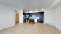 This single-floor space showcases a clean, modern design centered around an open-concept layout that seamlessly integrates the kitchen and living areas. The kitchen is a standout feature, outfitted with full-height dark navy cabinetry that provides ample storage and a bold contrast against the white walls and light hardwood-style flooring. A classic white subway tile backsplash adds texture and visual interest behind the countertops, while stainless steel appliances — including a refrigerator, range, and built-in microwave — offer a sleek, contemporary finish. A compact butcher-block island with a wooden base sits at the center of the kitchen, providing additional workspace and casual dining or prep space. Recessed lighting throughout the kitchen area ensures bright, even illumination, complementing the open and airy feel of the space. The light-toned flooring extends seamlessly throughout, creating a cohesive and spacious atmosphere. White walls and a textured ceiling contribute to the overall brightness of the interior. A doorway visible from the main area hints at additional rooms beyond, rounding out this well-appointed and functional living space.