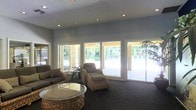This single-story space functions as a comfortable lounge or common area, designed to blur the boundary between interior and exterior environments. The most prominent architectural feature is the wall of large sliding glass doors and picture windows that span nearly the entire exterior-facing side of the room, flooding the interior with abundant natural light and offering unobstructed views of a landscaped outdoor courtyard. The courtyard beyond features mature trees, manicured shrubs, and a covered patio area with a ceiling fan, suggesting a well-maintained outdoor retreat.

Inside, the room is finished with dark wall-to-wall carpeting that grounds the space and provides a warm contrast to the light grey walls and white window frames. Recessed lighting in the ceiling supplements the generous natural light. The seating arrangement consists of wicker-style chairs with cushions and a matching wicker coffee table with a glass top, creating a casual and inviting atmosphere. A small indoor plant near the entrance adds a natural accent. The overall ambiance is relaxed and open, with an emphasis on connectivity to the outdoors.