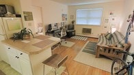 This single-floor space showcases an open-concept layout that seamlessly integrates a dining area, home office, and living room into one cohesive environment. Warm hardwood flooring runs throughout, lending a sense of continuity and natural warmth to the space. A breakfast bar with bar-height seating defines the dining area, offering a casual spot for meals or conversation.

The home office section is equipped with a dedicated workstation, ergonomic seating, and framed artwork that adds a personalized, lived-in feel. Adjacent to it, the living area is anchored by a window unit air conditioner and a large window fitted with horizontal blinds, allowing natural light to filter through while maintaining privacy. Striped and neutral-toned area rugs help delineate each functional zone within the open floor plan.

The living room accommodates a media setup alongside comfortable seating, with the overall palette kept light and neutral through cream-colored walls. Baseboard heating is visible along the perimeter, indicating a practical approach to climate control. The space presents a functional, well-organized layout suited for everyday living and working.