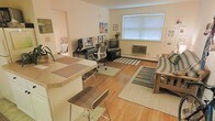 This single-floor space showcases an open-concept layout that seamlessly integrates a dining area, home office, and living room into one cohesive environment. Warm hardwood flooring runs throughout, lending a sense of continuity and natural warmth to the space. A breakfast bar with bar-height seating defines the dining area, offering a casual spot for meals or conversation.

The home office section is equipped with a dedicated workstation, ergonomic seating, and framed artwork that adds a personalized, lived-in feel. Adjacent to it, the living area is anchored by a window unit air conditioner and a large window fitted with horizontal blinds, allowing natural light to filter through while maintaining privacy. Striped and neutral-toned area rugs help delineate each functional zone within the open floor plan.

The living room accommodates a media setup alongside comfortable seating, with the overall palette kept light and neutral through cream-colored walls. Baseboard heating is visible along the perimeter, indicating a practical approach to climate control. The space presents a functional, well-organized layout suited for everyday living and working.