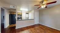 This single-floor unit is designed with a practical, open layout that connects the living area and kitchen seamlessly. Rich, warm-toned hardwood-style flooring runs throughout the space, creating a cohesive and inviting atmosphere. The living area is generously sized, featuring a ceiling fan with a light fixture and a sliding glass door that opens to a private outdoor patio, offering natural light and outdoor views of surrounding greenery.

The kitchen is positioned behind a peninsula-style breakfast bar topped with a dark countertop, which serves as both a functional workspace and a visual divider between the kitchen and living area. The kitchen is equipped with black appliances, including a refrigerator and an electric range, complemented by warm wood-toned cabinetry providing ample storage. A utility area adjacent to the kitchen houses a stacked washer and dryer unit, adding in-unit laundry convenience.

The bathroom features hardwood-style flooring consistent with the rest of the unit, a wood-toned vanity with a dark countertop, and a full tub-shower combination with white tile surround. Neutral wall tones and a textured ceiling are present throughout, contributing to a clean, understated aesthetic across the entire space.