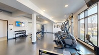 Located at 425 Ballantrae Drive in Winnipeg, this modern fitness facility offers a comprehensive workout environment across a generously sized single floor. The space features a clean, contemporary design with white walls, structural columns, and durable rubberized flooring suited to high-traffic exercise use. Recessed ceiling lighting throughout ensures the space is consistently well-lit, complemented by large floor-to-ceiling windows along one wall that flood the cardio zone with abundant natural light and provide views of the surrounding neighbourhood.

The main workout area is thoughtfully organized to accommodate a variety of training modalities. A dedicated cardio section lines the windowed wall and features multiple treadmills, elliptical trainers, and stair climbers, each equipped with individual mounted flat-screen monitors. A Concept2 RowERG rowing machine and a stationary bike are also available. The strength training zone includes a Hoist functional trainer cable machine, a weight bench, and a well-stocked dumbbell rack with a full range of free weights. A wall-mounted flat-screen TV adds an entertainment option for users. A large mirror in the strength area enhances the sense of space and supports proper form during exercise.

Additional amenities include a water fountain station, a seating bench, and access to change room facilities. The facility's layout ensures smooth circulation between zones, creating a practical and welcoming environment for fitness.