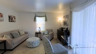 This single-story space presents a welcoming living area characterized by a neutral, understated color palette of soft grays and warm whites. The room is furnished with a comfortable sofa, multiple upholstered armchairs, and a patterned round ottoman that serves as a central accent piece, offering both style and versatility. A dark wood credenza along one wall provides functional storage and display space, topped with decorative items including candles and greenery. Wall art adds a subtle visual point of interest above the credenza. Natural light enters through a window dressed with elegant draped fabric valances in a muted sage green, complemented by blinds for light control. A floor lamp and a table lamp provide warm ambient lighting, enhancing the cozy atmosphere during evening hours. The light-toned carpeted flooring ties the room together, contributing to an overall sense of comfort and calm. A glass-top accent table near the entry adds a contemporary touch. The space is well-proportioned, offering enough room for relaxed seating arrangements while maintaining an open, uncluttered feel. The overall ambiance is calm, refined, and move-in ready.