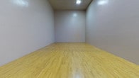 Located in Griffin, GA, this single-level racquetball court is a purpose-built athletic facility designed for competitive and recreational play. The court features a classic hardwood floor finished in a light natural tone, complete with clearly marked boundary and service lines that meet standard racquetball court specifications. The smooth, painted walls on all sides provide the consistent ball-rebound surface essential for gameplay, while the generous ceiling height allows for full, unrestricted play. Overhead fluorescent lighting ensures the court is evenly and adequately illuminated throughout. Entry to the court is provided through full-height glass doors, which also function as a viewing wall, allowing spectators to observe play from the adjacent area. A staircase with a metal railing leads to an elevated viewing gallery positioned above the entry, offering an excellent vantage point overlooking the entire court. An electrical panel is mounted on the wall near the entry. The overall design is clean, functional, and focused entirely on athletic performance, making this a well-equipped facility for racquetball enthusiasts in the Griffin community.