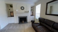 This single-floor space centers around a classic white-mantled fireplace framed by elegant crown molding and crisp white walls, creating a refined yet welcoming atmosphere. The fireplace surround features grey tile detailing and is flanked by decorative candleholders, adding a traditional charm to the room. A framed panel above the mantel serves as a focal point, complemented by tasteful décor including a round wall clock and small accent pieces.

To the right of the fireplace, built-in shelving and cabinetry provide both display space and functional storage, with a dark countertop surface offering a practical workspace or display area. The cabinetry extends downward with white lower cabinets, maintaining a cohesive, clean aesthetic. A window fitted with white horizontal blinds allows natural light to filter into the room, brightening the neutral-toned carpet that runs throughout.

The seating area features a dark brown leather sofa accented with a floral throw pillow, positioned to take advantage of both the fireplace and the natural light. Overall, the space conveys a comfortable, traditionally styled environment with thoughtful built-in details and a warm, neutral palette.