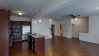 This two-story residence presents an open-concept main level that seamlessly integrates the kitchen and living areas into one expansive, cohesive space. Rich, dark hardwood flooring extends throughout, adding warmth and continuity to the layout.

The kitchen is well-appointed with dark wood cabinetry, stainless steel appliances including a full-size refrigerator, and a central island featuring granite countertops and a built-in sink. Two pendant lights hang above the island, providing focused task lighting that complements the recessed lighting overhead.

The open living area flows naturally from the kitchen, offering generous room to arrange furnishings comfortably. A ceiling fan with a light fixture serves the space, contributing to both comfort and illumination. Multiple interior doors on the main level suggest access to additional rooms, including what appears to be a bathroom.

A staircase with a wood railing leads to the upper floor, which likely contains additional living or sleeping quarters. The neutral white walls throughout the main level provide a clean, versatile backdrop, making the space feel bright and adaptable to a variety of interior styles.