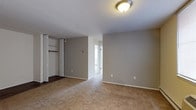 This single-floor residential space offers a clean and functional layout with a straightforward, practical design. The bedroom is a standout feature, showcasing wall-to-wall carpeting in a warm beige tone that adds comfort underfoot. Neutral gray walls create a calm, versatile backdrop that pairs well with a variety of décor styles. A built-in closet with bifold mirrored doors provides convenient storage, featuring interior shelving and a hanging rod for organized use of space. A flush-mount ceiling light fixture provides overhead illumination, contributing to the room's simple yet tidy aesthetic. White baseboards frame the perimeter of the room, offering a crisp contrast to the soft gray walls. A doorway visible from within the bedroom leads to an adjacent area, suggesting a connected and accessible floor plan. The space benefits from natural light filtering in from a window visible through the doorway, lending a sense of openness and brightness. Overall, this space presents a no-frills, move-in-ready interior well-suited for comfortable everyday living.
