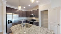 Located at 20 Pine Hall Dr in Clayton, NC, this single-story home offers a thoughtfully designed layout with modern finishes throughout. The kitchen serves as a central highlight, showcasing rich dark-espresso cabinetry extending to the ceiling, light granite countertops, and a full suite of stainless steel appliances including a French door refrigerator, electric range, and built-in microwave. A large island with an undermount sink and pendant lighting adds both functionality and style, while a dedicated walk-in pantry with built-in shelving provides generous storage. Light wood-look flooring runs through the main living areas, complementing the kitchen's warm, contemporary aesthetic. The primary bedroom features plush carpeting, a tray ceiling with crown molding detail, a ceiling fan, and a window overlooking the exterior. Additional bedrooms are carpeted and include ceiling fans, with one featuring notably elevated ceilings. Bathrooms are finished with granite-topped dark vanities and light wood-look tile flooring, each equipped with a tub-shower combination featuring tiled surrounds and built-in shelving. Walk-in closets with wire shelving systems offer ample wardrobe storage. Crown molding and recessed lighting are consistent throughout, lending a polished, cohesive character to the entire home.