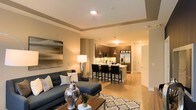 This contemporary single-floor residence showcases a thoughtfully designed open-concept layout that effortlessly blends living, dining, and kitchen spaces into one cohesive environment. The living area is anchored by a plush dark sectional sofa, complemented by a patterned area rug and a wooden coffee table, creating a warm yet sophisticated gathering space. A large abstract wall art piece adds a bold visual focal point to the room. Warm recessed lighting and an elegant tray ceiling lend architectural depth and a refined ambiance throughout.

The kitchen is a standout feature, boasting rich dark wood cabinetry, stainless steel appliances — including a French door refrigerator and built-in microwave — and a spacious island with bar seating for four. Sleek pendant lights hang above the island, adding a modern industrial touch that ties the space together beautifully.

Light wood-toned flooring runs throughout, enhancing the sense of openness and continuity. Neutral warm tones on the walls and ceiling create an inviting atmosphere, while ample recessed lighting ensures the space is bright and welcoming. A hallway leading to additional rooms is visible, hinting at further well-appointed spaces beyond the main living area.