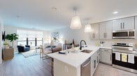 This thoughtfully designed single-floor residence showcases a bright, open-concept layout that seamlessly connects the kitchen, dining, and living areas. The kitchen is a standout feature, centered around a large island with a white quartz countertop, undermount sink, and matte black fixtures. Gray shaker-style cabinetry with black hardware, a classic subway tile backsplash, and a full suite of stainless steel appliances — including a range, over-the-range microwave, and refrigerator — complete this well-equipped space. Pendant lighting adds warmth above the island. The adjoining dining area flows naturally into a comfortable living room, which is bathed in natural light from large windows with roller shades offering views of the surrounding area. Light wood-tone flooring runs throughout the main living spaces, adding warmth and continuity. The bedroom is a serene retreat, featuring large windows dressed in sheer curtains that invite abundant natural light. The bathroom is finished with a walk-in glass-enclosed shower, a gray vanity with a white countertop, a large mirror, and clean tile flooring, delivering a polished, modern aesthetic. The overall ambiance is fresh, contemporary, and welcoming.