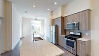 This single-floor residence offers a thoughtfully designed open-concept layout that seamlessly connects the kitchen and living areas. The kitchen is a standout feature, showcasing a large granite-topped island with a double basin sink, ideal for both meal preparation and casual dining. Warm, wood-toned cabinetry pairs elegantly with a tile backsplash, while stainless steel appliances — including a gas range, over-the-range microwave, and refrigerator — provide a sleek, modern finish. Recessed lighting throughout the kitchen ensures the space is bright and well-illuminated at all times. The open layout flows naturally toward the living area, where a ceiling fan and a large window bathe the space in natural light and offer a pleasant view of surrounding greenery. Wood-style flooring runs throughout, adding warmth and continuity across the living and kitchen spaces. Clean white walls and a neutral color palette create a calm, inviting ambiance that is both contemporary and versatile. This well-appointed residence combines functional design with modern aesthetics, making it a comfortable and stylish place to call home.