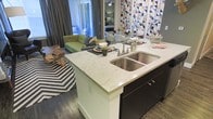 This single-floor residence showcases a thoughtfully designed open-concept layout that effortlessly connects the kitchen, dining, and living areas into one cohesive and inviting space. At the heart of the kitchen stands a striking island topped with veined marble countertops, housing a deep double-basin stainless steel sink and offering generous bar seating — ideal for casual dining and entertaining. Dark espresso cabinetry provides a bold contrast to the light marble surfaces, while rich hardwood-style flooring runs throughout, grounding the space with warmth and continuity.

The living area features floor-to-ceiling drapery that hints at expansive windows and access to an outdoor area, flooding the interior with natural light. A vibrant geometric accent wall anchors the dining zone, adding an artistic focal point that energizes the space. Eclectic furnishings, including a plush green sofa, a graphic chevron area rug, and a live-edge wood accent table, lend the space a curated, contemporary character.

The open sightlines create a sense of spaciousness, making the layout feel both functional and welcoming. The combination of modern finishes, bold design choices, and seamless flow between zones makes this a dynamic and well-appointed living environment.