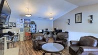 This single-floor space combines a relaxed lounge area with a functional workstation zone, creating a versatile environment suited for both leisure and productivity. The main area features rich hardwood-style flooring that runs throughout, complementing the warm, neutral-toned walls accented by classic white wainscoting panels along the lower half.

The lounge section is furnished with dark brown leather armchairs with nail-head trim detailing, arranged around a round glass-top coffee table, offering a welcoming and comfortable seating arrangement. Two pendant light fixtures hang from the smooth ceiling, providing warm, ambient illumination across the space.

Toward the back, a dedicated workstation area includes multiple computer desks with monitors and ergonomic office chairs, making it a practical workspace. A large bookshelf unit nearby stores books, binders, and decorative items, while colorful wall posters add a lively touch. A vintage-style popcorn cart positioned near the shelving adds a unique, charming character to the space. The overall ambiance is inviting and functional, blending comfort with practicality.