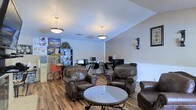This single-floor space combines a relaxed lounge area with a functional workstation zone, creating a versatile environment suited for both leisure and productivity. The main area features rich hardwood-style flooring that runs throughout, complementing the warm, neutral-toned walls accented by classic white wainscoting panels along the lower half.

The lounge section is furnished with dark brown leather armchairs with nail-head trim detailing, arranged around a round glass-top coffee table, offering a welcoming and comfortable seating arrangement. Two pendant light fixtures hang from the smooth ceiling, providing warm, ambient illumination across the space.

Toward the back, a dedicated workstation area includes multiple computer desks with monitors and ergonomic office chairs, making it a practical workspace. A large bookshelf unit nearby stores books, binders, and decorative items, while colorful wall posters add a lively touch. A vintage-style popcorn cart positioned near the shelving adds a unique, charming character to the space. The overall ambiance is inviting and functional, blending comfort with practicality.