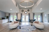 Located at 3822 Mizell Rd in Greensboro, NC, this single-story community space offers a thoughtfully designed, open-concept layout that seamlessly blends comfort and functionality. The interior is defined by light hardwood flooring, crisp white walls, and soaring ceilings that enhance the sense of spaciousness throughout.

The main gathering area serves as the heart of the space, anchored by a striking dark fireplace surround with a mounted flat-screen TV above. Clustered blue velvet accent chairs and a low oval coffee table create an inviting lounge setting, while large abstract artwork adds a contemporary artistic touch to the walls.

Adjacent to the lounge, a dedicated workspace features a sleek wooden desk and chair positioned beside a row of tall, narrow black-framed windows that flood the area with natural light and outdoor views. This area is well-suited for productivity in a refined setting.

The central reception area commands attention with a dramatic circular gold pendant light fixture that hangs from a raised ceiling. A curved cream chaise lounge, area rugs, and side tables contribute to an elegant yet welcoming atmosphere. A staff workstation and French glass doors provide a seamless transition between areas.

On the opposite end, French doors open to an outdoor patio, while a coffee station and additional seating areas with bistro-style tables complete this versatile, well-appointed community hub.