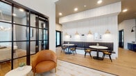 This thoughtfully designed single-floor space combines modern aesthetics with functional versatility. The entry area welcomes visitors with a geometric-patterned tile floor and curved cognac leather lounge chairs arranged around a round white side table. Bold black steel-frame glass doors provide a striking architectural detail while offering visual connectivity between zones, maintaining an open yet defined feel throughout.

A built-in banquette dining area features channel-tufted gray upholstered seating along a white shiplap accent wall, complemented by round tables, black Windsor-style chairs, and warm wood pendant lights. Framed artwork accents the wall above, adding a curated touch to the space.

The coffee bar area is equipped with deep navy shaker-style cabinetry topped with a concrete-finish countertop, a floating natural wood shelf, and white subway tile backsplash. Black sconce lighting and recessed ceiling lights illuminate the area effectively. Light oak hardwood flooring flows throughout much of the space, providing warmth and continuity.

A private dining or conference room features a large natural wood table, upholstered chairs, tall windows with white drapes, gray walls, a wood-planked ceiling, and a statement branch-style chandelier, creating an intimate yet refined atmosphere.