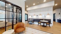 This thoughtfully designed single-floor space combines modern aesthetics with functional versatility. The entry area welcomes visitors with a geometric-patterned tile floor and curved cognac leather lounge chairs arranged around a round white side table. Bold black steel-frame glass doors provide a striking architectural detail while offering visual connectivity between zones, maintaining an open yet defined feel throughout.

A built-in banquette dining area features channel-tufted gray upholstered seating along a white shiplap accent wall, complemented by round tables, black Windsor-style chairs, and warm wood pendant lights. Framed artwork accents the wall above, adding a curated touch to the space.

The coffee bar area is equipped with deep navy shaker-style cabinetry topped with a concrete-finish countertop, a floating natural wood shelf, and white subway tile backsplash. Black sconce lighting and recessed ceiling lights illuminate the area effectively. Light oak hardwood flooring flows throughout much of the space, providing warmth and continuity.

A private dining or conference room features a large natural wood table, upholstered chairs, tall windows with white drapes, gray walls, a wood-planked ceiling, and a statement branch-style chandelier, creating an intimate yet refined atmosphere.