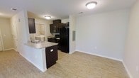 This single-floor space offers a clean, modern layout centered around an open-concept kitchen and living area. The kitchen is outfitted with dark espresso cabinetry that provides a bold contrast against the light-toned walls and hardwood-style flooring that runs throughout the unit. A peninsula with a granite-style countertop and an integrated sink serves as both a functional workspace and a natural divider between the kitchen and the living area, making the space feel connected yet purposefully organized. Black appliances — including a refrigerator, gas range, and built-in microwave — lend a cohesive, contemporary aesthetic to the kitchen. The open living area is bright and airy, with neutral white walls and clean white baseboards that enhance the sense of openness. Recessed flush-mount ceiling lighting provides consistent illumination across the space. A utility area with a washer and dryer is visible just off the kitchen, adding everyday convenience. The overall ambiance is modern and functional, with a neutral palette that offers a versatile backdrop for a variety of interior styles.
