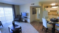 This single-floor space presents an open-concept layout that seamlessly connects the living area to the dining space and beyond. The living room is carpeted and furnished with a patterned accent chair, a flat-screen television on a dark wood console table, and a low-profile coffee table, creating a comfortable and functional arrangement. Neutral-toned walls throughout lend a clean, understated aesthetic that allows the furnishings to stand out.

A transition from carpet to hardwood-style flooring marks the shift toward the dining area, where a white high-back chair is visible alongside what appears to be a bar-height table or counter. A decorative triptych wall art piece and a pair of decorative vases on a shelf add visual interest near the entryway to the hallway.

The hallway connects the main living space to additional rooms, with natural light visible at the far end, suggesting well-lit secondary spaces. An overhead HVAC vent is centrally located in the ceiling, and a textured ceiling finish is consistent throughout. The overall ambiance is modest, tidy, and move-in ready, offering a practical and cohesive living environment.