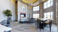 This single-story space makes a striking first impression with its dramatic double-height ceilings, which lend an expansive, open feel to the interior. A central tray ceiling, finished in a warm golden yellow and accented with recessed lighting, draws the eye upward and frames a pair of large, globe-shaped crystal chandeliers that serve as dazzling focal points. Towering floor-to-ceiling windows line one wall, flooding the space with abundant natural light and offering views of the surrounding landscape, complemented by flowing floor-length drapery.

The living area centers around a floor-to-ceiling brick fireplace surround, framed by white cabinetry and topped with a flat-screen television—creating a polished, functional focal point. A sectional sofa in a neutral gray anchors this lounge area, paired with a leather ottoman and additional seating. Adjacent to this is a high-top bar or communal table accompanied by tall upholstered chairs, making it well-suited for casual dining or collaborative use. The patterned carpeted flooring adds texture and visual interest throughout, tying the various seating zones together into a cohesive, sophisticated communal environment.