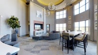 This single-story space makes a striking first impression with its dramatic double-height ceilings, which lend an expansive, open feel to the interior. A central tray ceiling, finished in a warm golden yellow and accented with recessed lighting, draws the eye upward and frames a pair of large, globe-shaped crystal chandeliers that serve as dazzling focal points. Towering floor-to-ceiling windows line one wall, flooding the space with abundant natural light and offering views of the surrounding landscape, complemented by flowing floor-length drapery.

The living area centers around a floor-to-ceiling brick fireplace surround, framed by white cabinetry and topped with a flat-screen television—creating a polished, functional focal point. A sectional sofa in a neutral gray anchors this lounge area, paired with a leather ottoman and additional seating. Adjacent to this is a high-top bar or communal table accompanied by tall upholstered chairs, making it well-suited for casual dining or collaborative use. The patterned carpeted flooring adds texture and visual interest throughout, tying the various seating zones together into a cohesive, sophisticated communal environment.