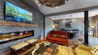 Located at 4125 Hayden Lofts Place in Dublin, Ohio, this single-floor community clubhouse showcases a bold industrial-chic aesthetic that blends rustic materials with modern finishes throughout. The main lounge area serves as the centerpiece of the space, anchored by a dramatic floor-to-ceiling stacked stone accent wall housing a sleek linear fireplace with a reclaimed wood mantel. A large wall-mounted television sits above, while a distinctive gear-shaped ceiling fixture with integrated lighting adds an industrial focal point overhead. Warm hardwood flooring runs throughout, complementing the earthy tones of the furnishings, including a vibrant red sofa, woven area rugs, and unique natural wood coffee tables. A secondary seating area features soaring ceilings, exposed metal sliding barn-style doors with iron rod detailing, pendant Edison bulb lighting, and additional lounge seating for a relaxed, loft-inspired atmosphere. The dining area is appointed with round stone-top tables and wicker seating beneath industrial cage pendant lights. Adjacent to the dining space, a well-equipped kitchenette offers dark granite countertops, stainless steel cabinetry, a sink, microwave, and additional appliances — ideal for gatherings and events. Bold "HL" lettering on the wall reinforces the Hayden Lofts brand identity throughout.