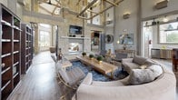 This single-floor clubhouse-style space is designed to impress, combining rustic warmth with modern sophistication throughout its distinct zones. The centerpiece is a grand lounge area anchored by a floor-to-ceiling natural stone fireplace, above which a large flat-screen television is mounted. Soaring ceilings with exposed wooden beam accents and geometric gold-toned chandeliers draw the eye upward, while hardwood flooring and a rich blue area rug ground the seating arrangement of neutral-toned sofas, tufted ottomans, and accent chairs. Tall dark bookshelves with decorative accessories line the walls, adding both function and character.

Adjacent to the lounge, a coffee and tea bar features a reclaimed wood island base topped with white quartz countertops, a patterned tile backsplash, and large windows that invite natural light and outdoor views. Upholstered navy blue barstools with nailhead trim complete the inviting nook.

The games room offers another dramatic voluminous ceiling and expansive grid-pane windows overlooking the surrounding property. This area includes a pool table, shuffleboard table, and leather seating, with vintage-style décor adding a playful, social atmosphere throughout.